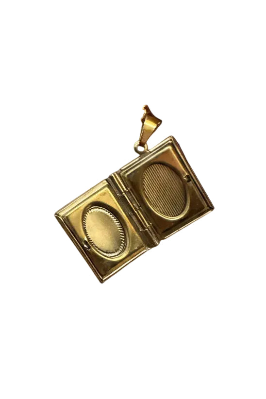 A gold-toned square locket pendant with two oval compartments, held open by a hand against a textured brick wall background.
