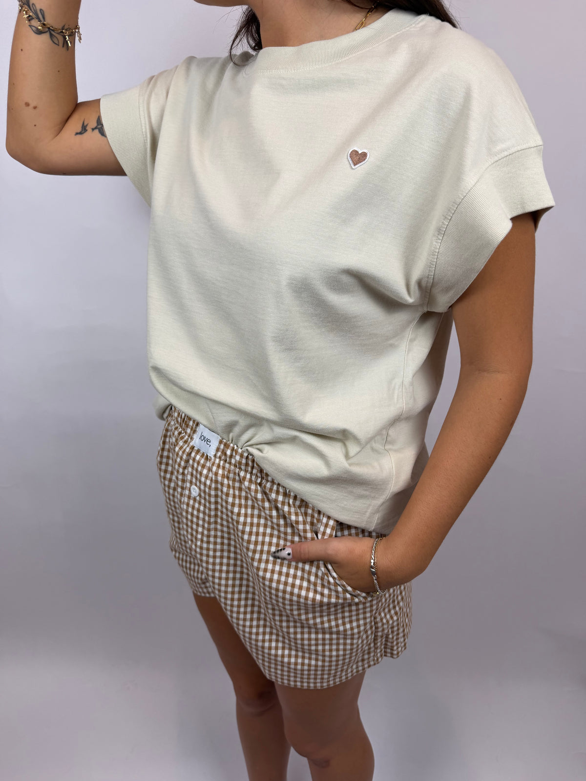 A person wearing a cream-colored short-sleeve t-shirt with a small embroidered heart on the chest, paired with brown and white gingham shorts, against a plain light gray background.