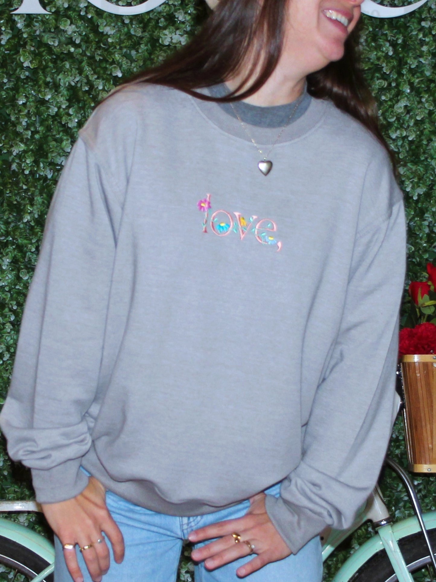 A person wearing a gray crewneck sweatshirt with a floral logo embroidered on the chest in multiple colors.