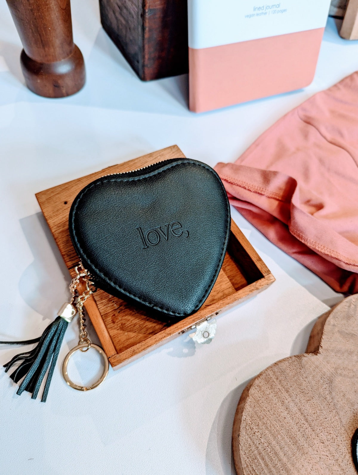 A heart-shaped black vegan leather wallet with the word 'love' embossed on it, displayed on a wooden surface. It includes a gold keyring and a matching tassel.