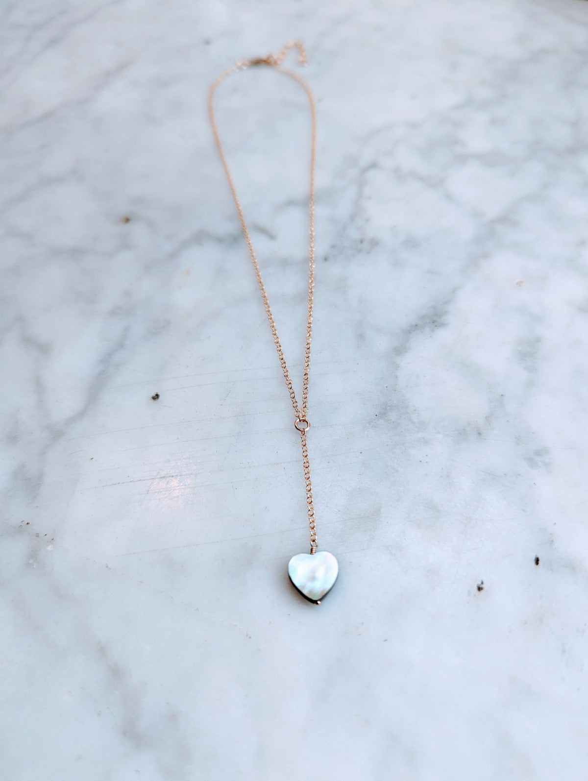 A smoky grey heart-shaped seashell lariat necklace on a 14k gold-filled chain, displayed on a marble surface.