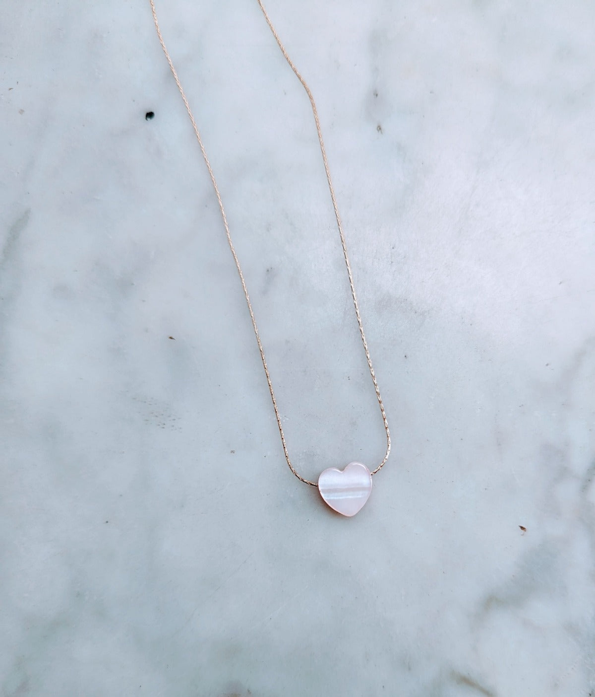 A pale pink heart-shaped seashell necklace on a 14k gold-filled chain, displayed on a marble surface.