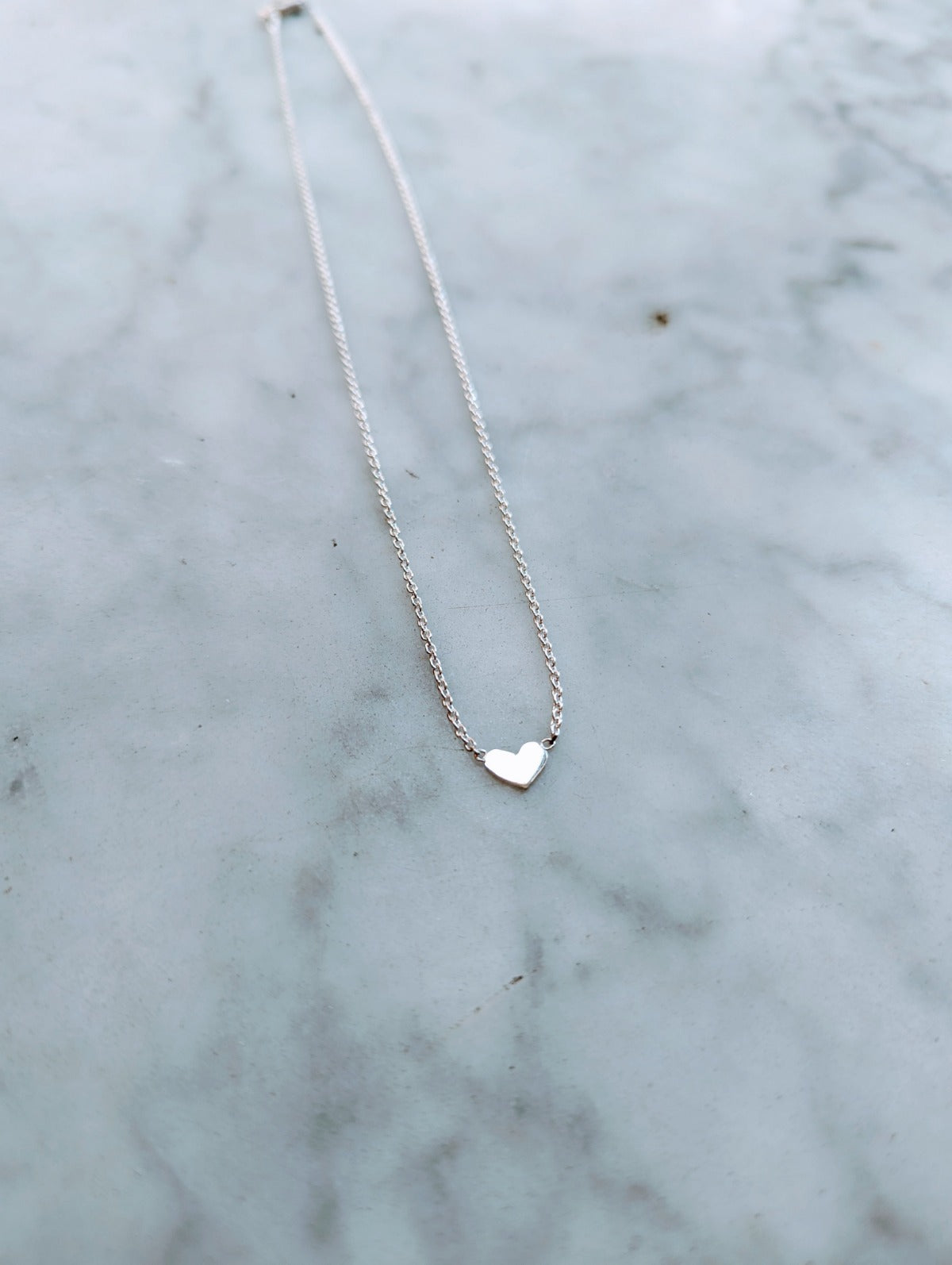 A small heart-shaped sterling silver necklace on a delicate chain, displayed on a marble surface.