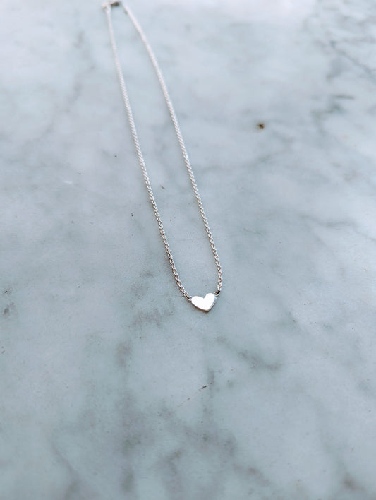 A small heart-shaped sterling silver necklace on a delicate chain, displayed on a marble surface.