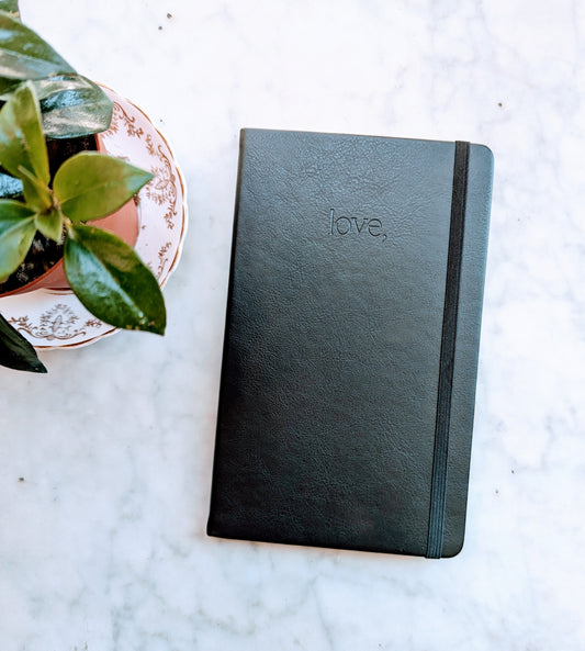 A black vegan leather cover journal with the word 'love' debossed on the front, placed on a marble surface next to a plant.