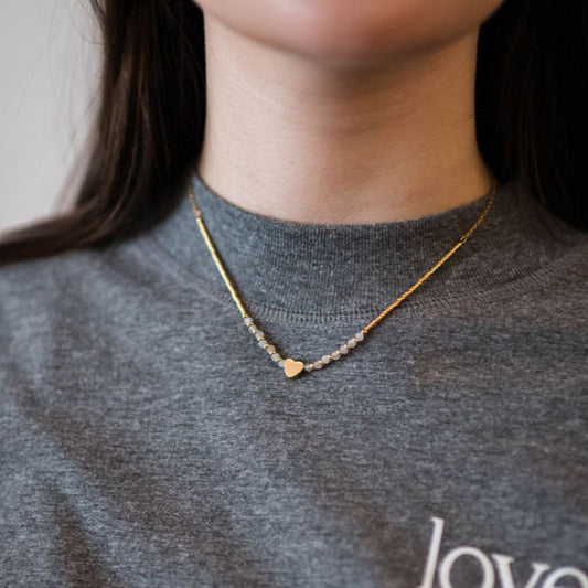 A person wearing a grey sweater and a gold-plated stainless steel chain necklace with a heart-shaped bead and quartz stones.