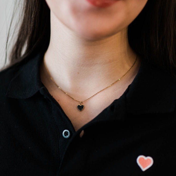 A person wearing a black shirt with a gold-plated sterling silver necklace featuring a midnight black enamel heart charm.