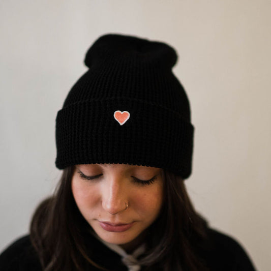 A person wearing a black knit beanie with a pink embroidered heart patch on the front cuff.