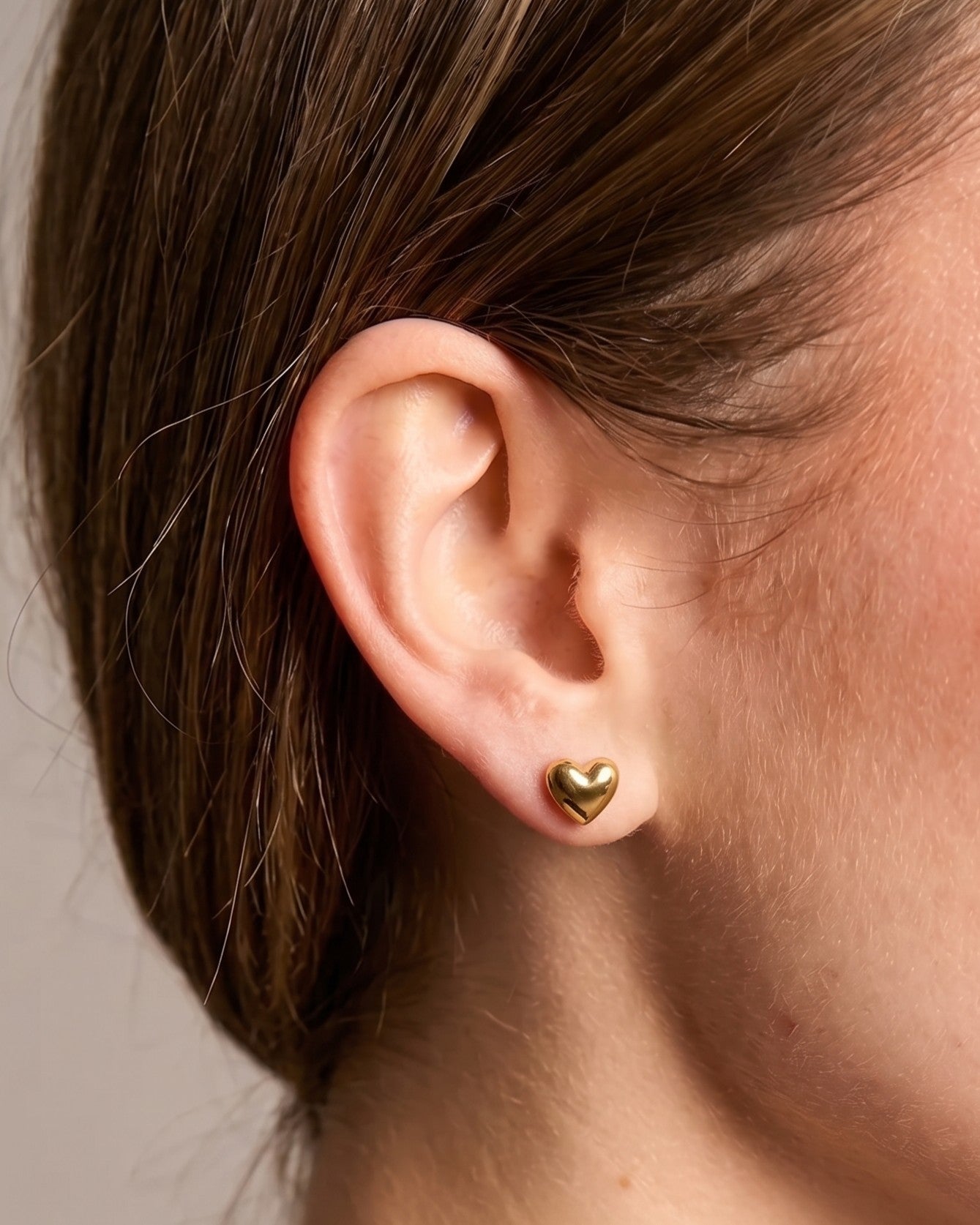 A close-up of a person's ear wearing a small polished gold heart stud earring against a soft neutral background.