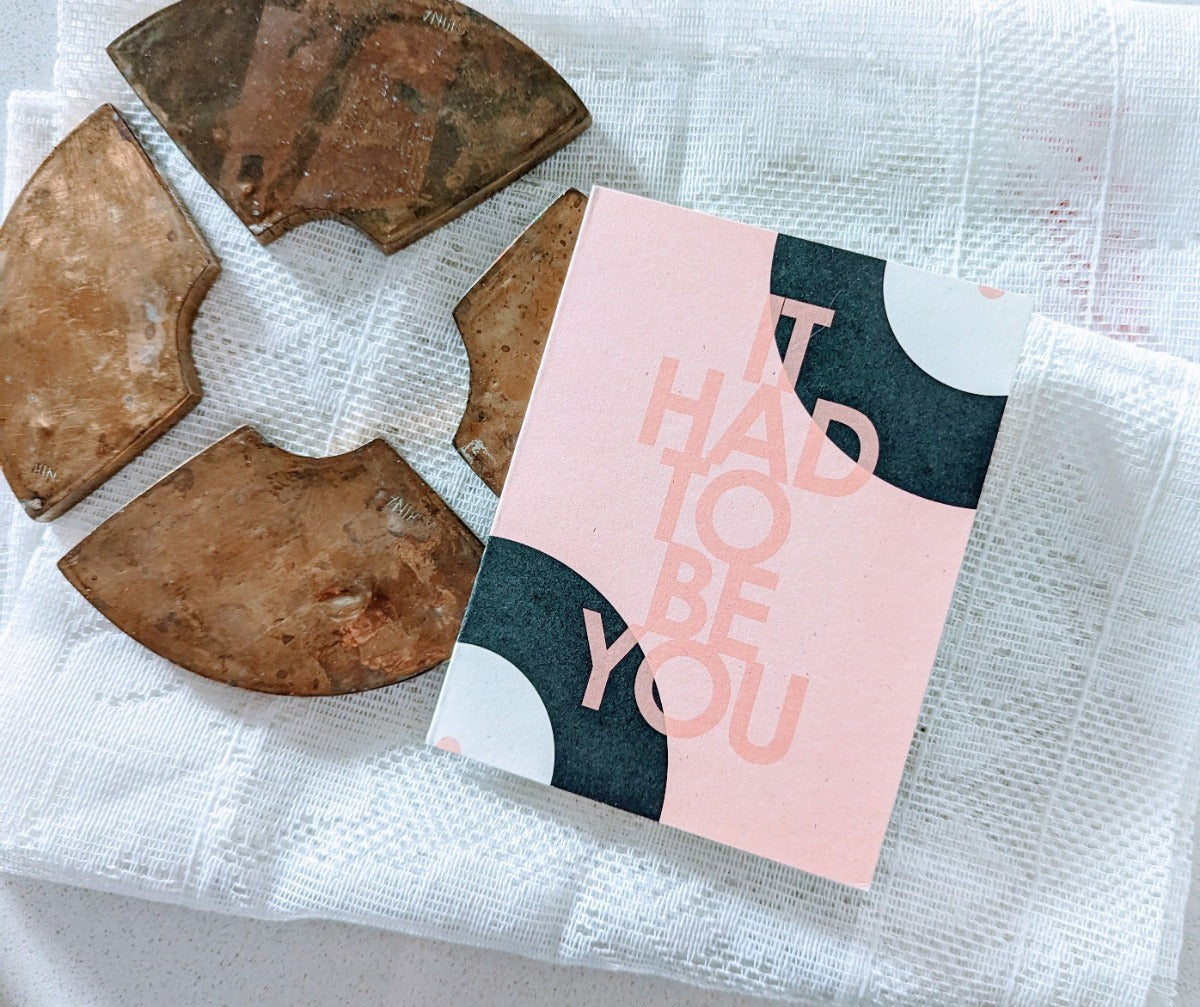 A greeting card with the phrase 'IT HAD TO BE YOU' printed in pink letters, accompanied by a matching envelope, displayed on a white surface with an abstract pattern.
