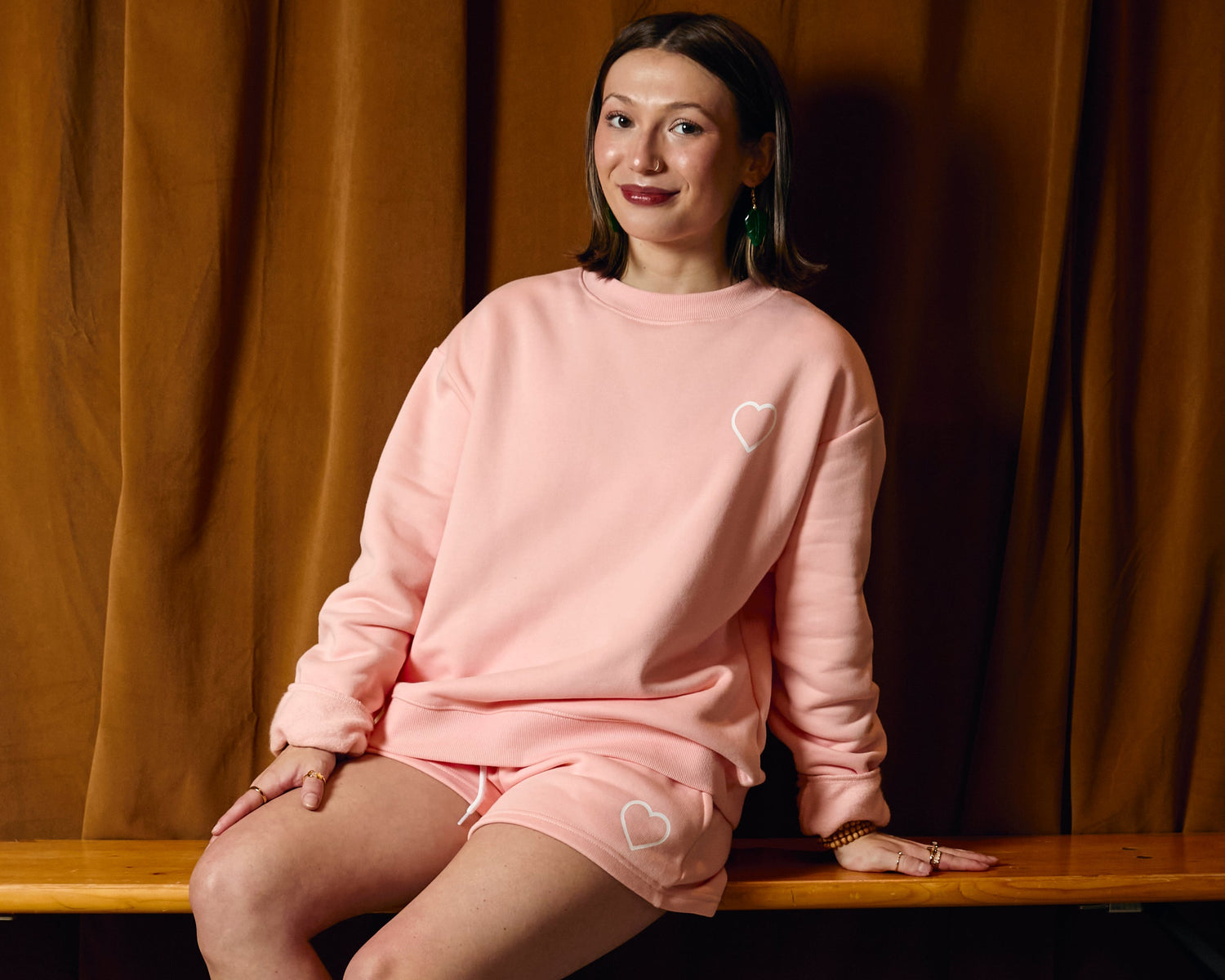 Woman in pink sweatshirt and shorts sitting on a bench against a brown curtain.
