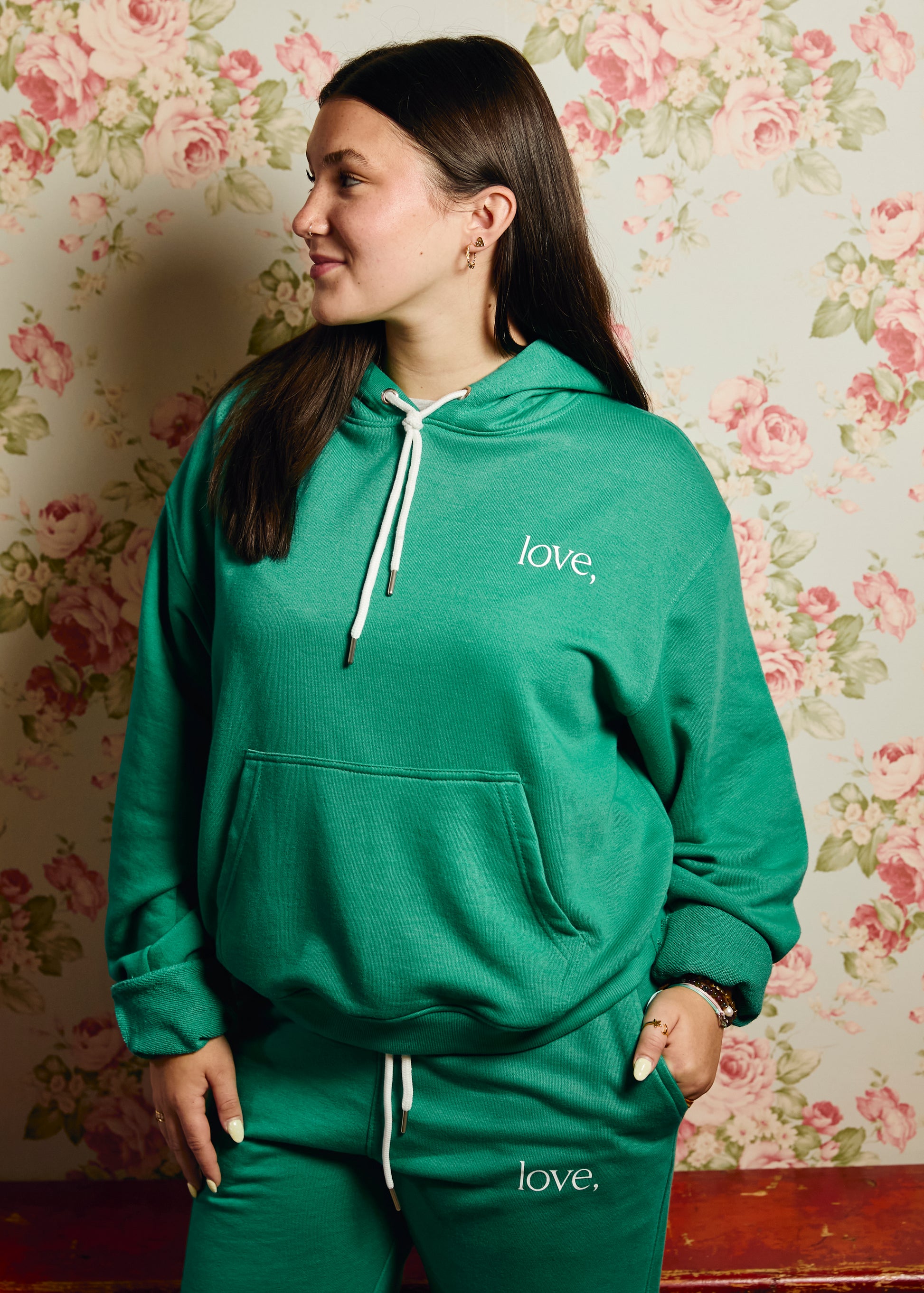 Woman wearing a green 'love' hoodie and pants against a floral wallpaper background