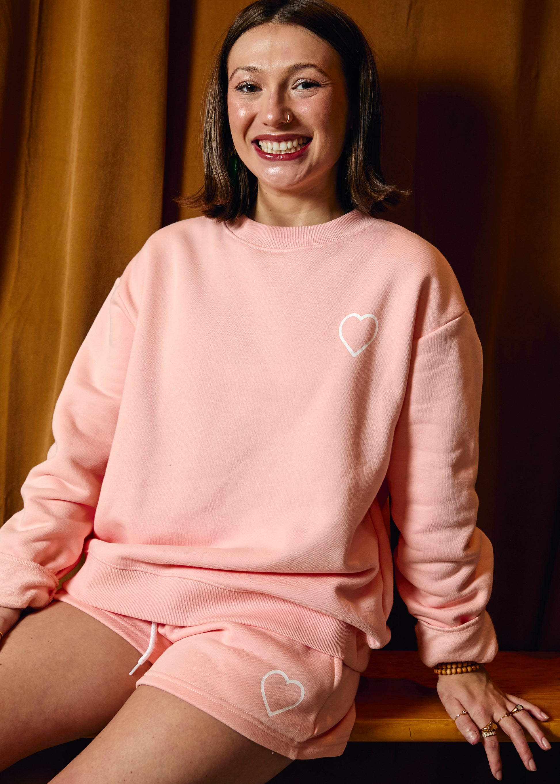Woman wearing a pink sweatshirt and shorts set with heart designs, sitting against a warm-toned curtain.