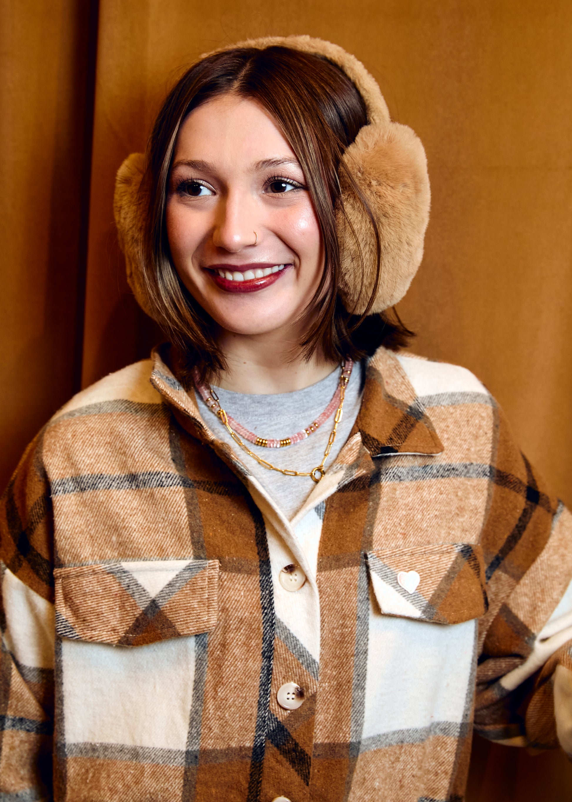 Person wearing a plaid coat and beige earmuffs against a brown background