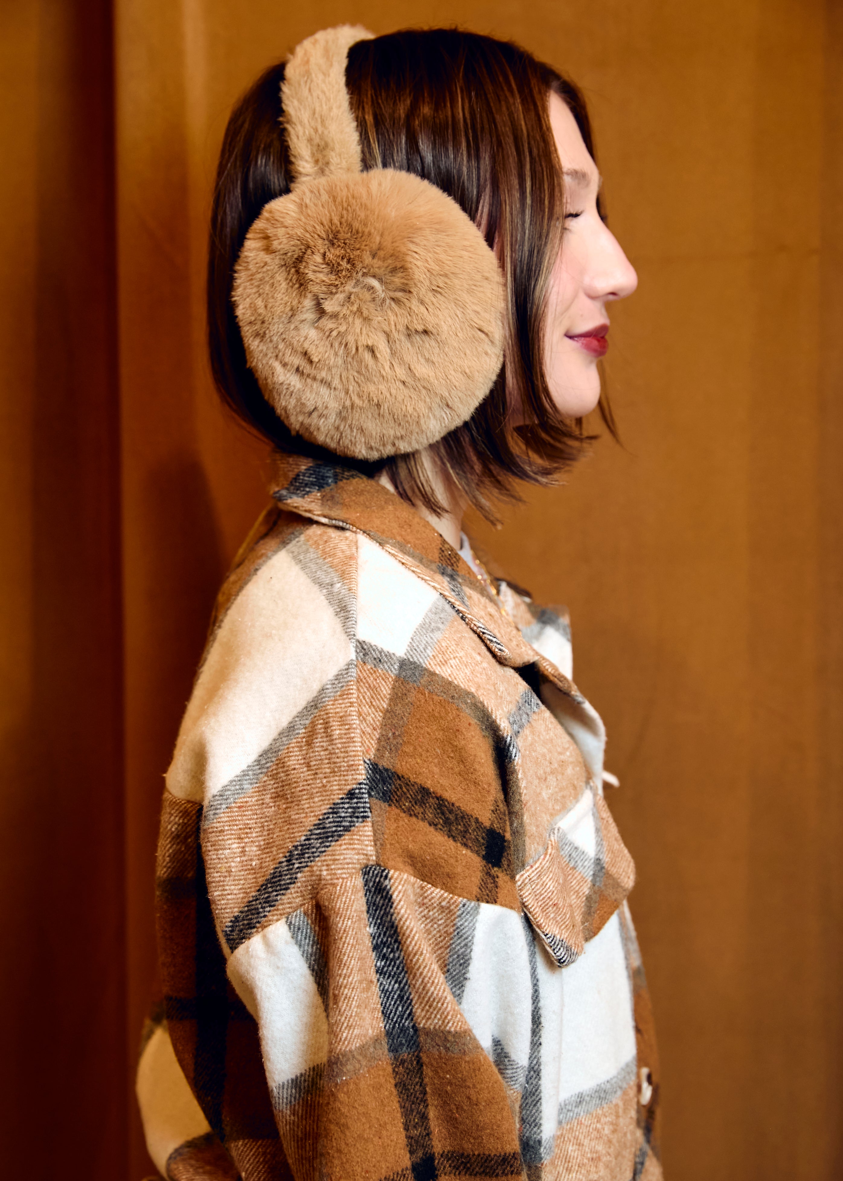 Person wearing beige earmuffs and a plaid coat against a brown background