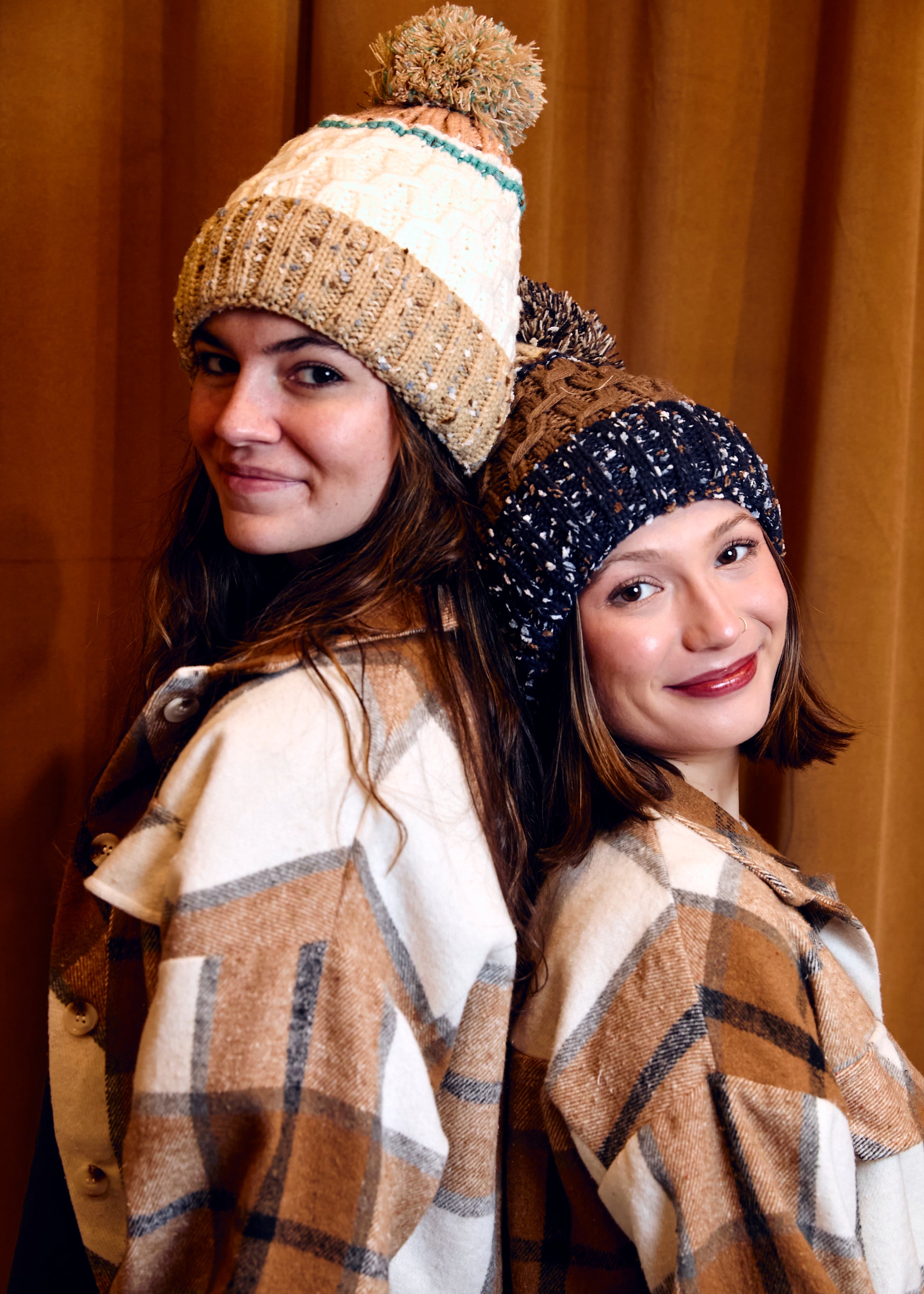 Two women wearing winter hats and plaid scarves against a brown background