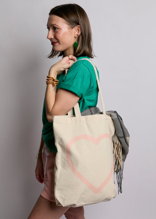 Woman holding a beige tote bag with a pink heart design against a plain background