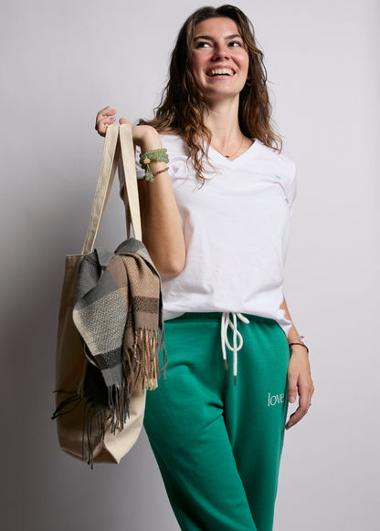 Woman holding a beige tote bag with a gray scarf, wearing a white shirt and green pants, on a plain background.