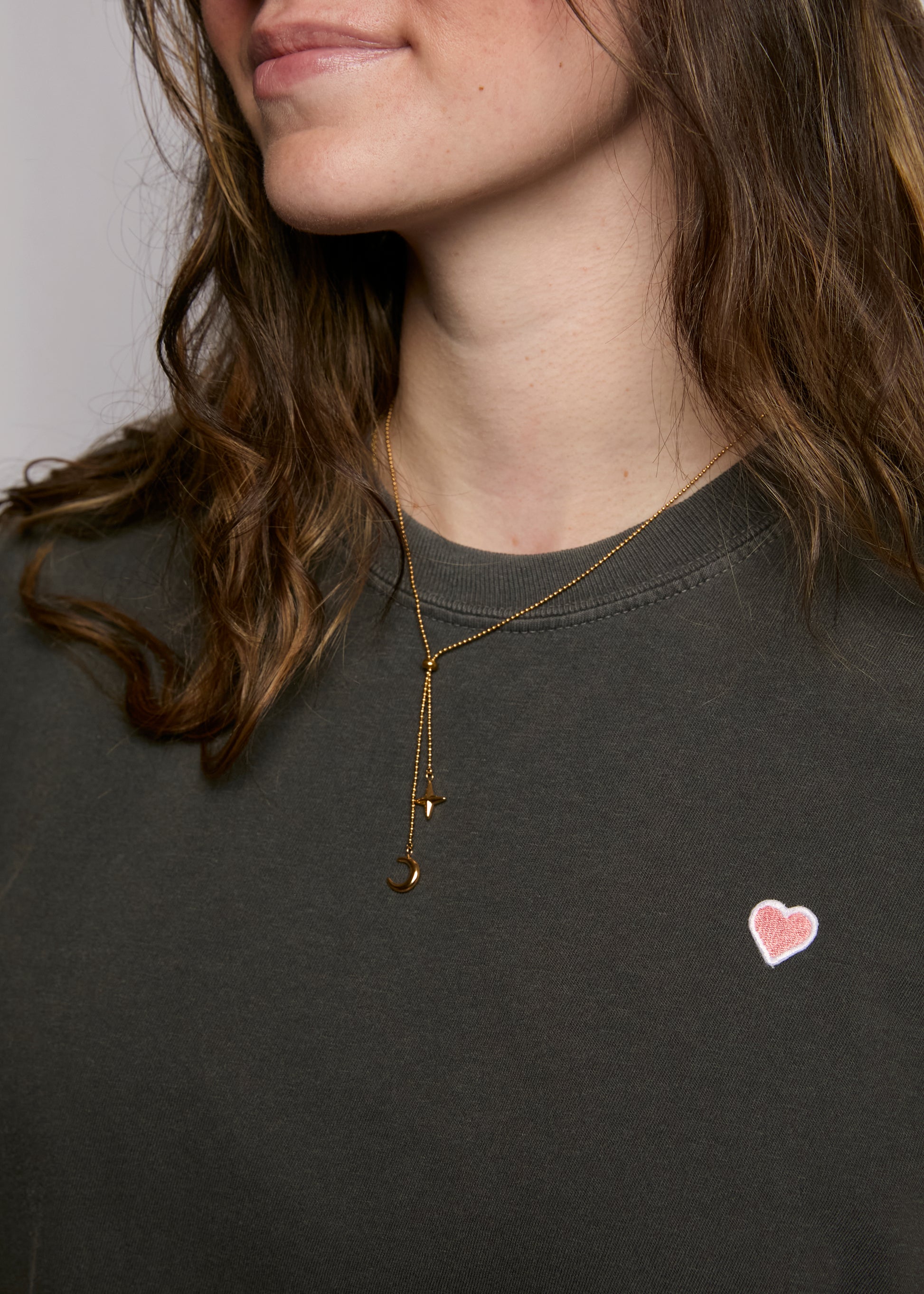 Woman wearing a gold necklace with a heart pendant on a dark shirt.
