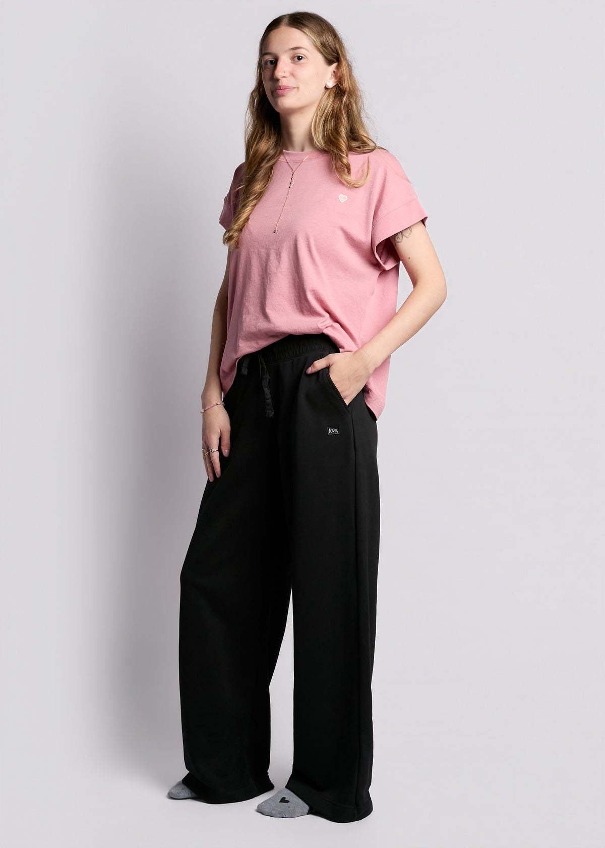 young woman wearing loose fit short sleeve pink tee with embroidered pink heart on left chest with loose fit black sweatpants with rectangle "love"" patch on left hip, with grey heather socks with black hearts on feet.