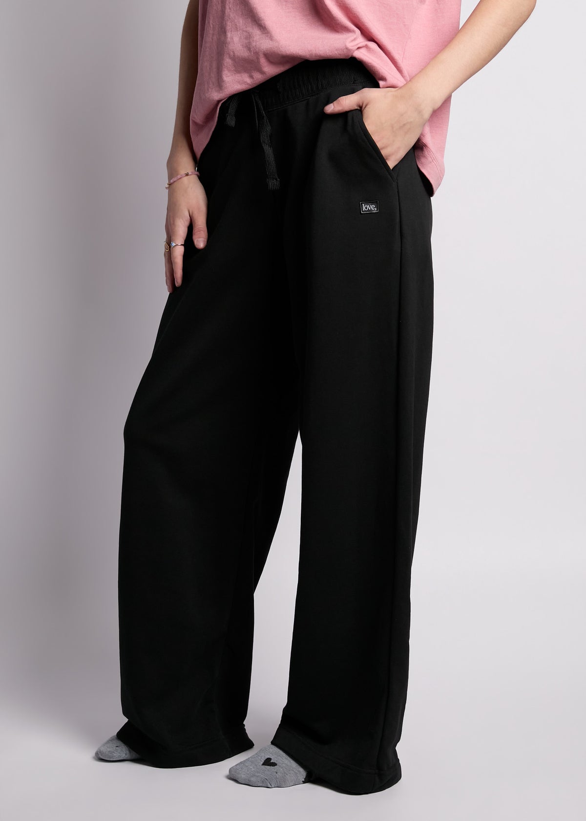 young woman wearing loose fit short sleeve pink tee with embroidered pink heart on left chest with loose fit black sweatpants with rectangle "love"" patch on left hip, with grey heather socks with black hearts on feet.