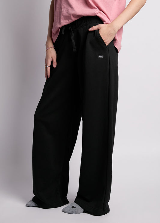 young woman wearing loose fit short sleeve pink tee with embroidered pink heart on left chest with loose fit black sweatpants with rectangle "love"" patch on left hip, with grey heather socks with black hearts on feet.