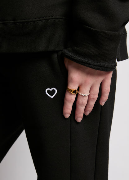 Person wearing black pants with a heart design, holding hands with another person on a light gray background.