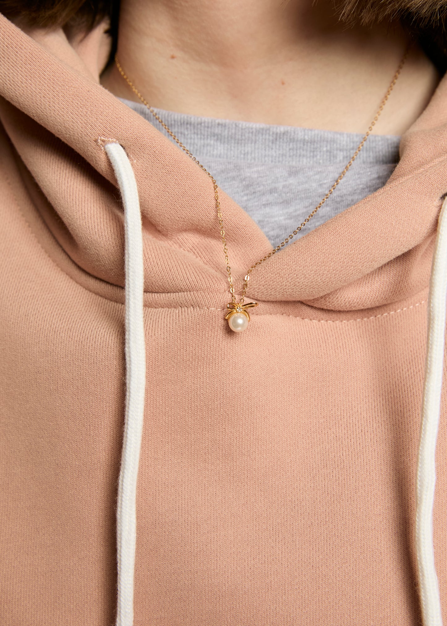 Close-up of a person wearing a beige hoodie with a necklace featuring a small pendant.