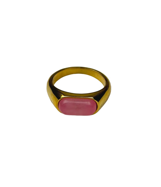 A gold signet ring with an oval pink cabochon gemstone, displayed on a light gray textured surface.