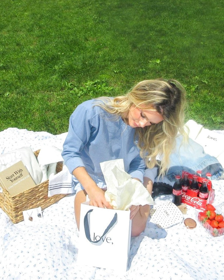 Woman sitting on a picnic blanket with a basket, gift bag, and drinks on a grassy field.