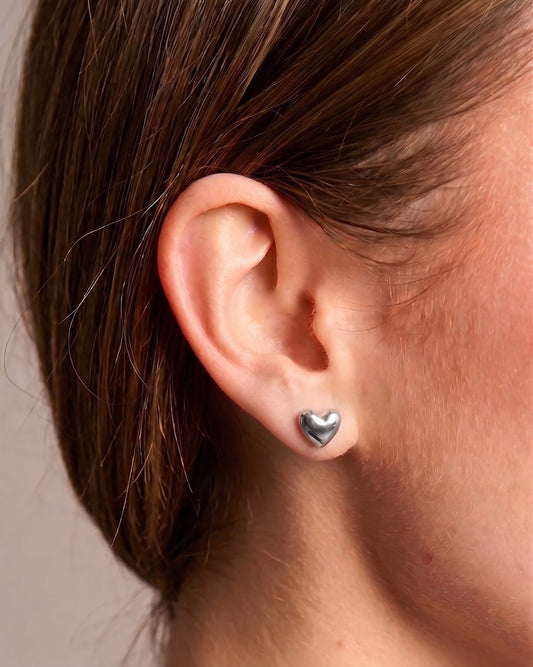 A close-up of a person's ear wearing a small polished silver heart stud earring.