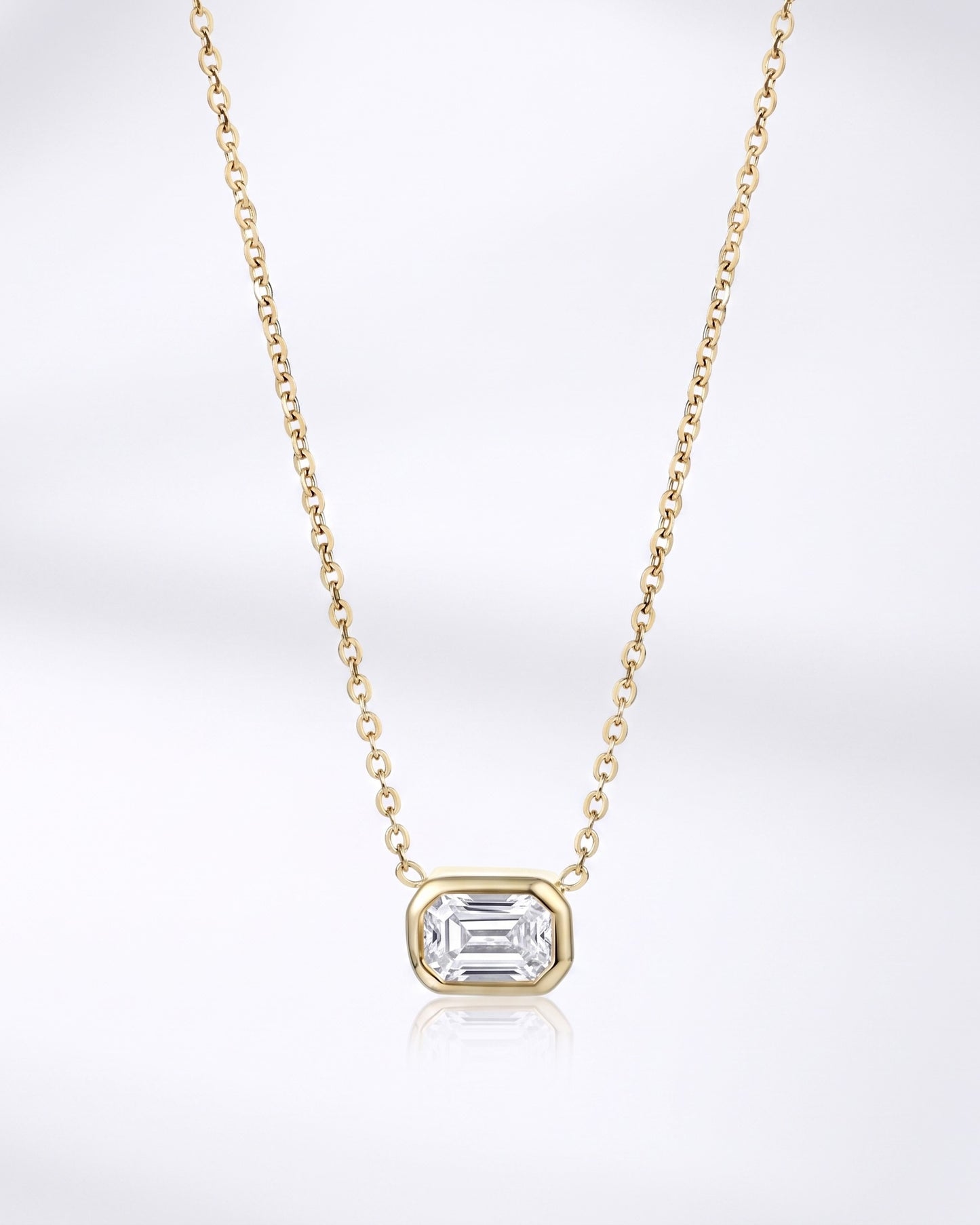 A delicate yellow gold chain necklace featuring a small rectangular bezel-set clear gemstone pendant, displayed against a clean white background with a soft reflection below.