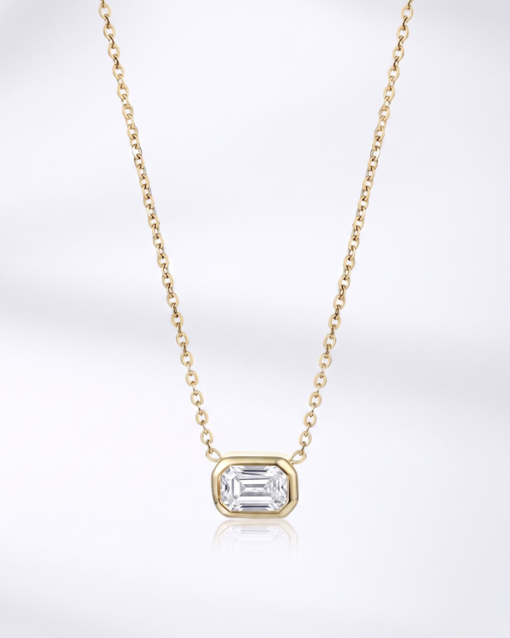 A delicate yellow gold chain necklace featuring a small rectangular bezel-set clear gemstone pendant, displayed against a clean white background with a soft reflection below.