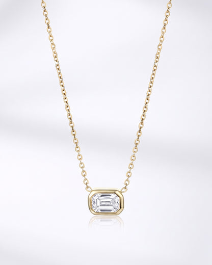 A delicate yellow gold chain necklace featuring a small rectangular bezel-set clear gemstone pendant, displayed against a clean white background with a soft reflection below.