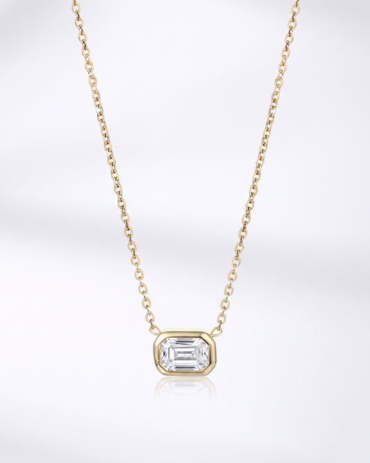 A delicate yellow gold chain necklace featuring a small rectangular bezel-set clear gemstone pendant, displayed against a clean white background with a soft reflection below.
