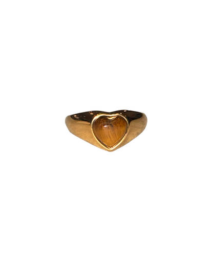 A gold signet ring with a heart-shaped face, featuring a warm brown tiger's eye inlay, resting on a textured light gray surface.