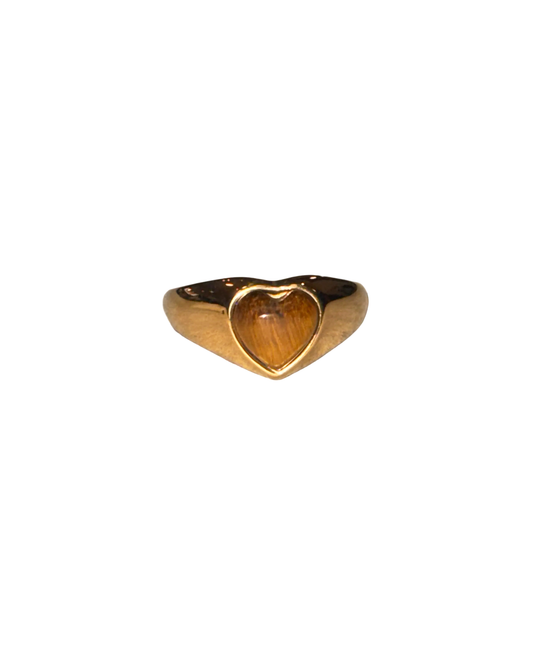 A gold signet ring with a heart-shaped face, featuring a warm brown tiger's eye inlay, resting on a textured light gray surface.